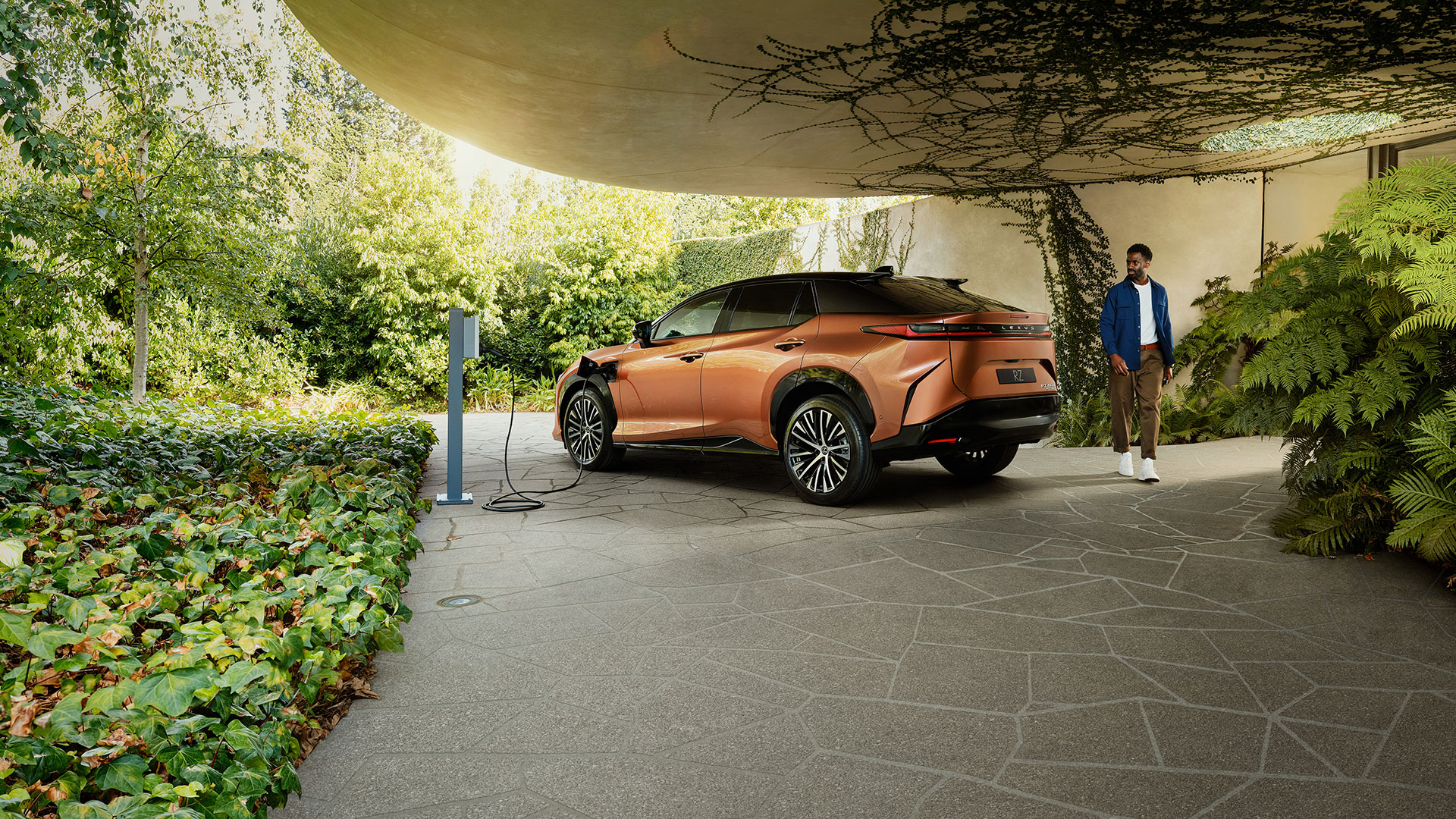 An orange Lexus RZ is parked and charging in a leafy green, vine-filled driveway. A man is seen walking around the rear of the car.