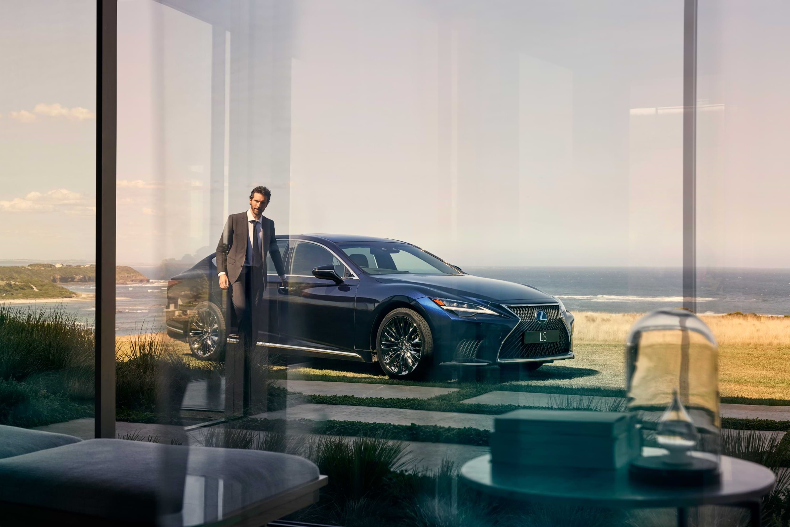 Man appears through a large glass window, hand on the handle of the driver's door to a Lexus LS hybrid.