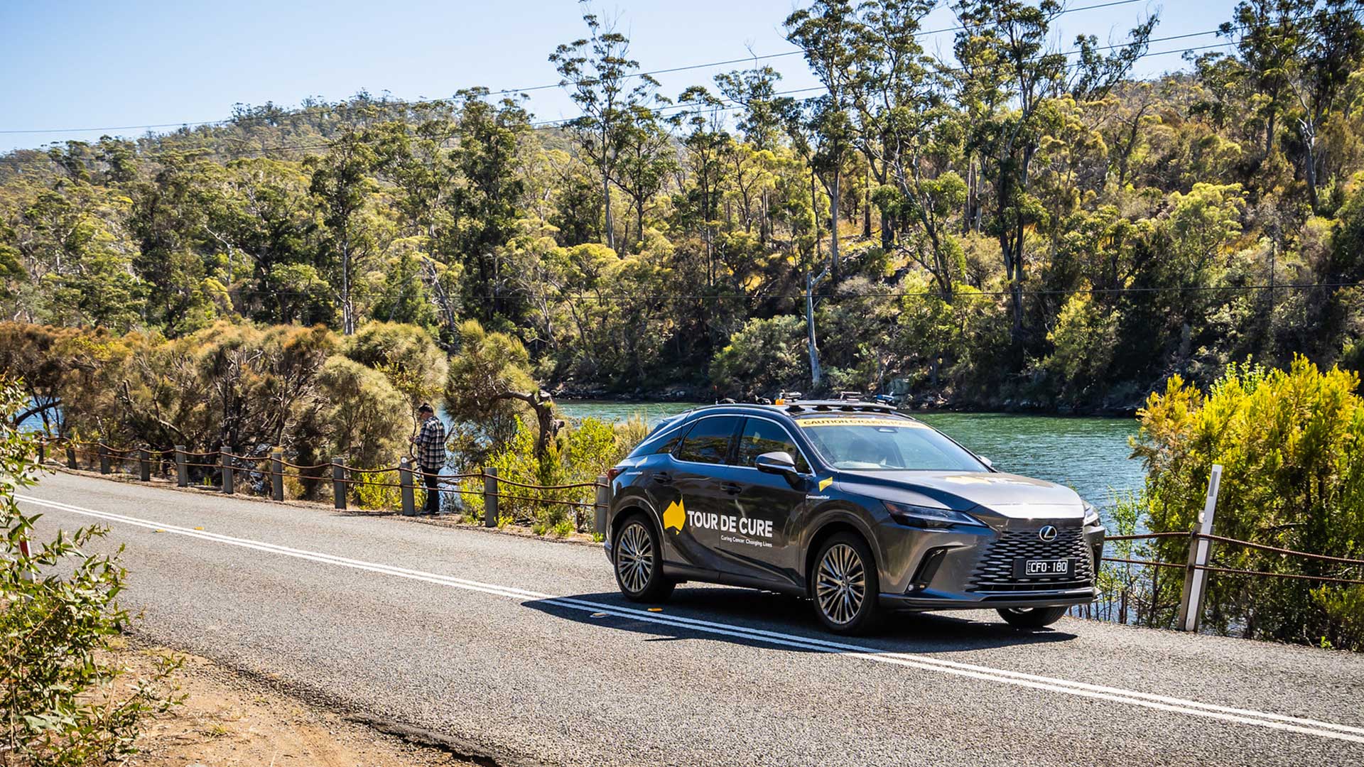 A Lexus Tour de Cure support vehicle drives along a road beside a river.