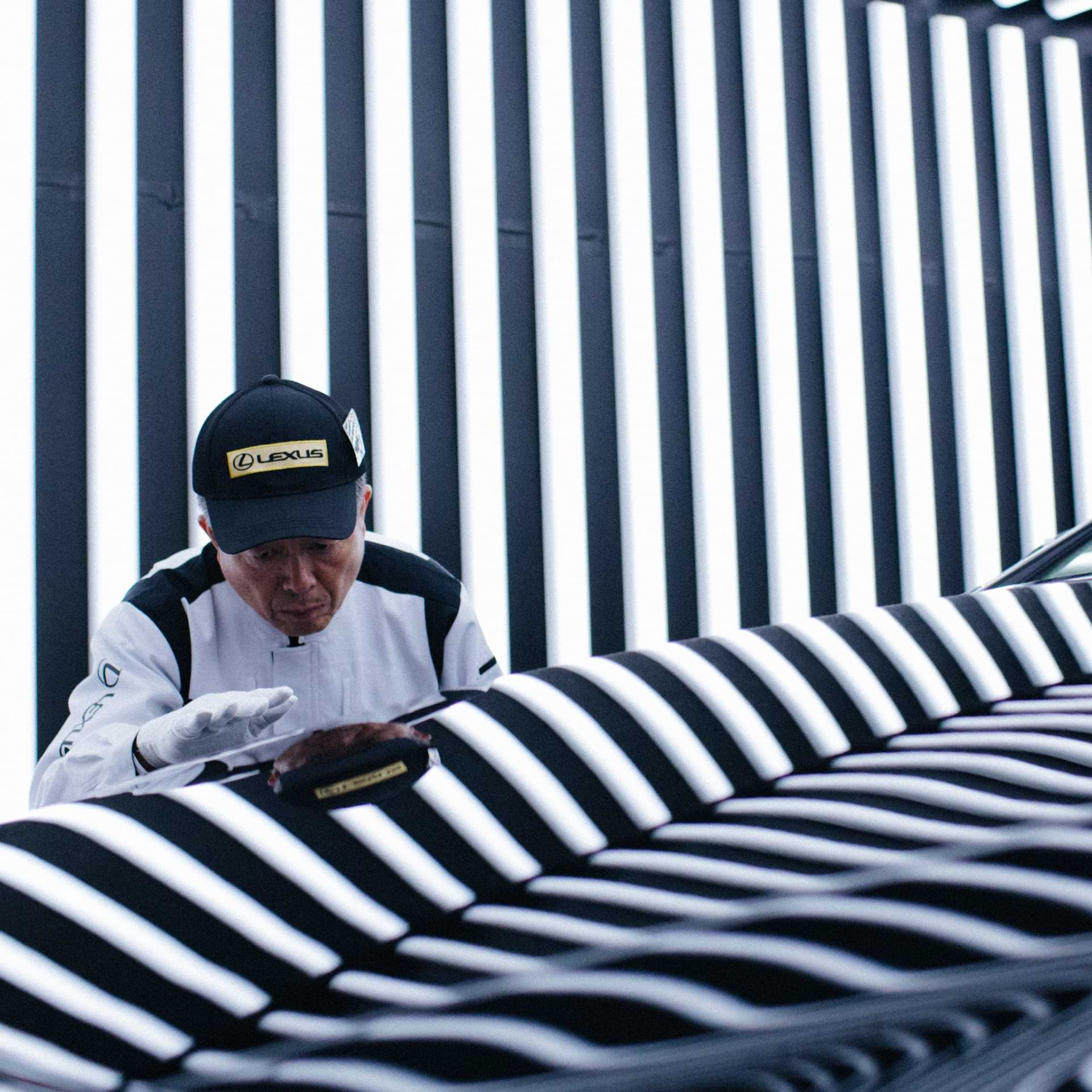 Lexus Takumi craftsman inspecting a vehicle surface under striped lighting inside a production facility.