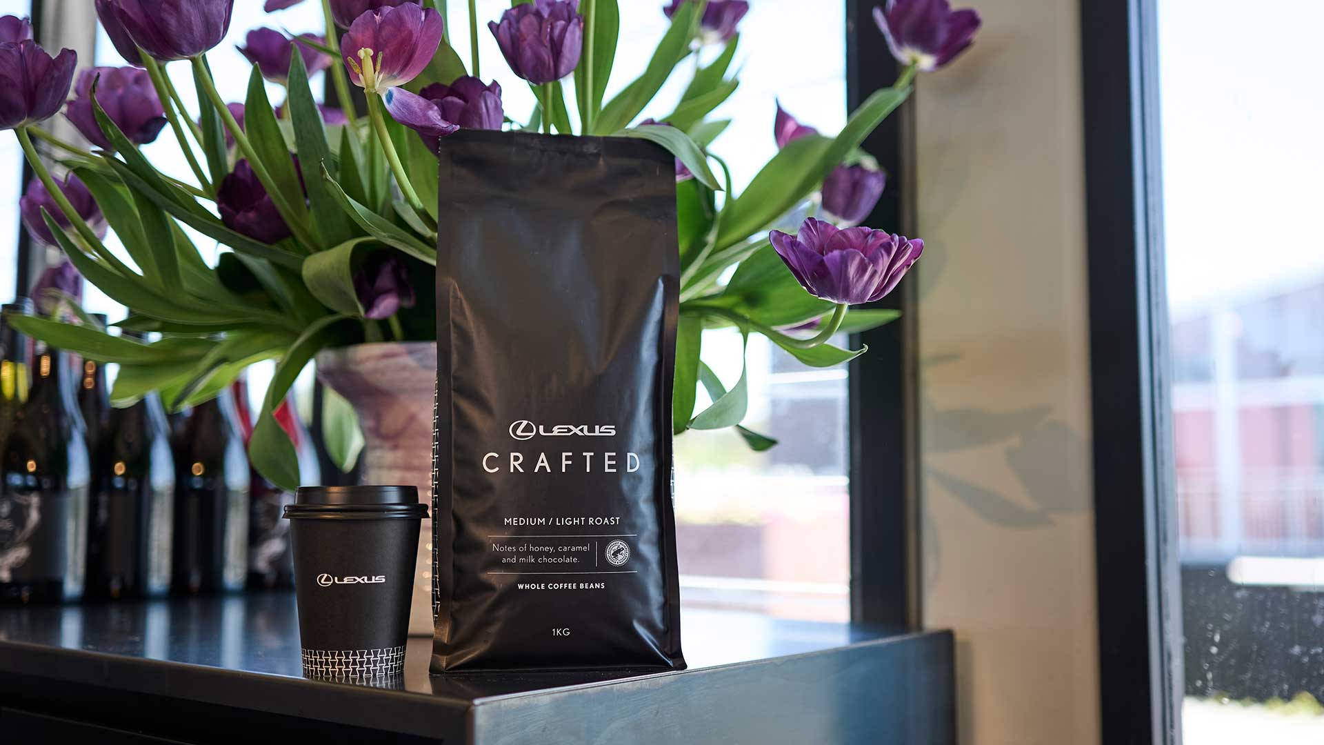 A bag of Lexus coffee beans sits on a counter with purple tulips in the background