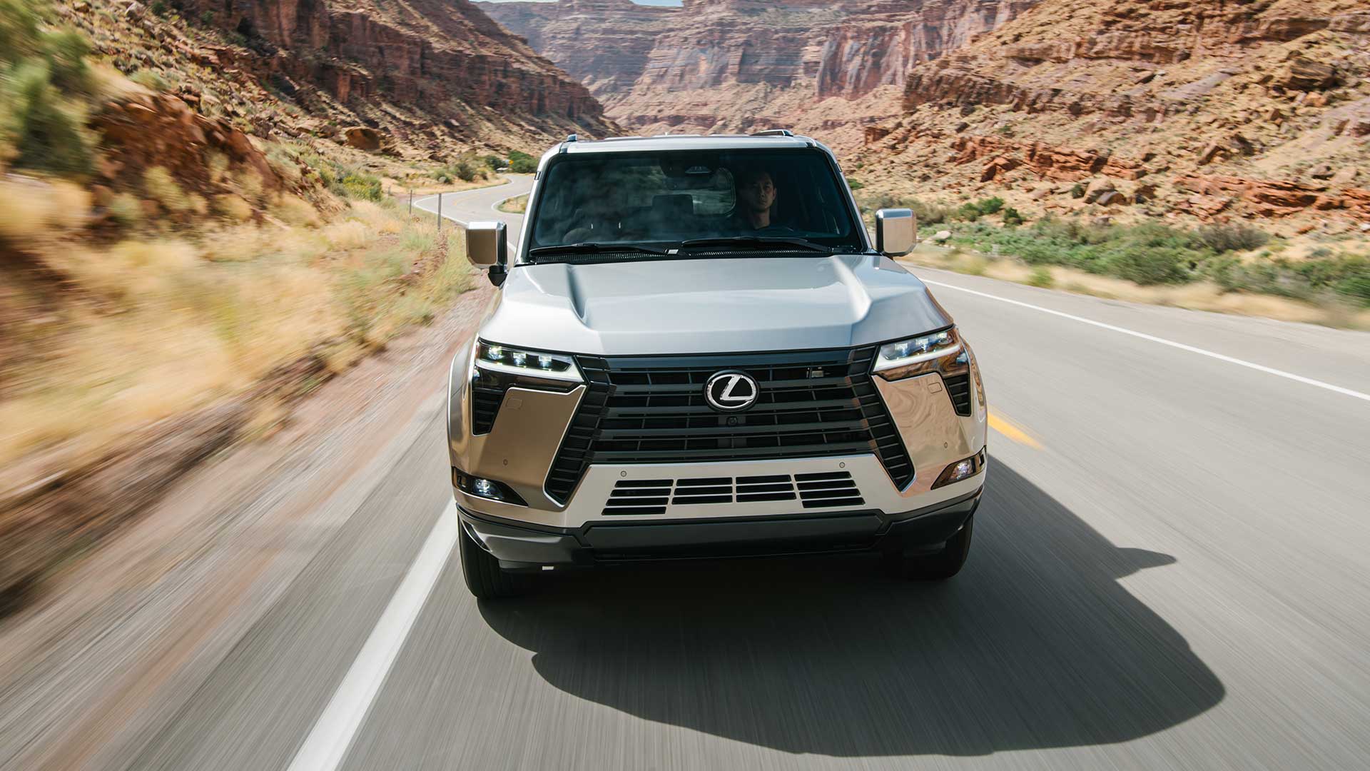 The front of a silver Lexus GX driving on a road with mountains in the background