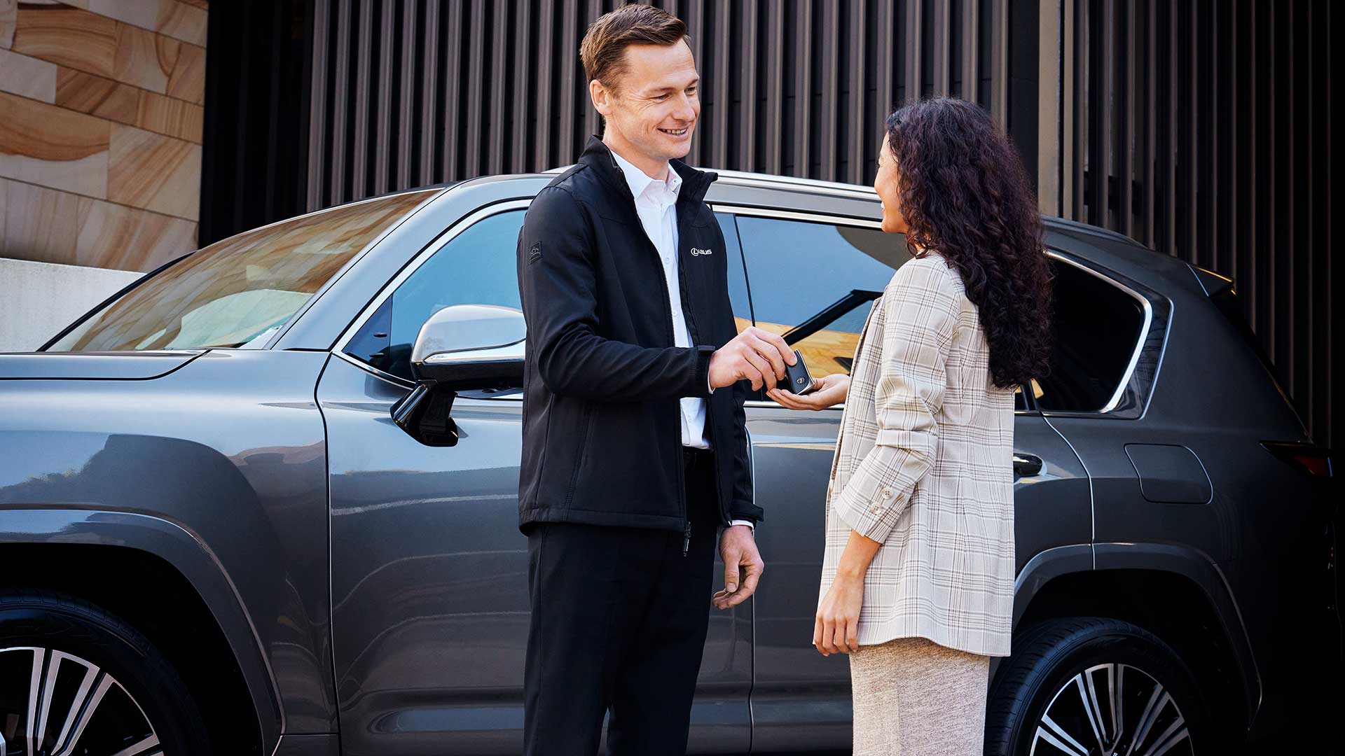 A man handing a woman keys in front of a grey Lexus vehicle
