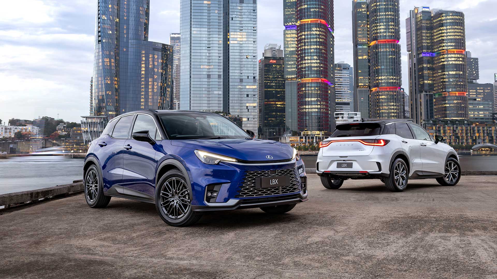 Two Lexus LBX vehicles, blue and white, parked against a cityscape