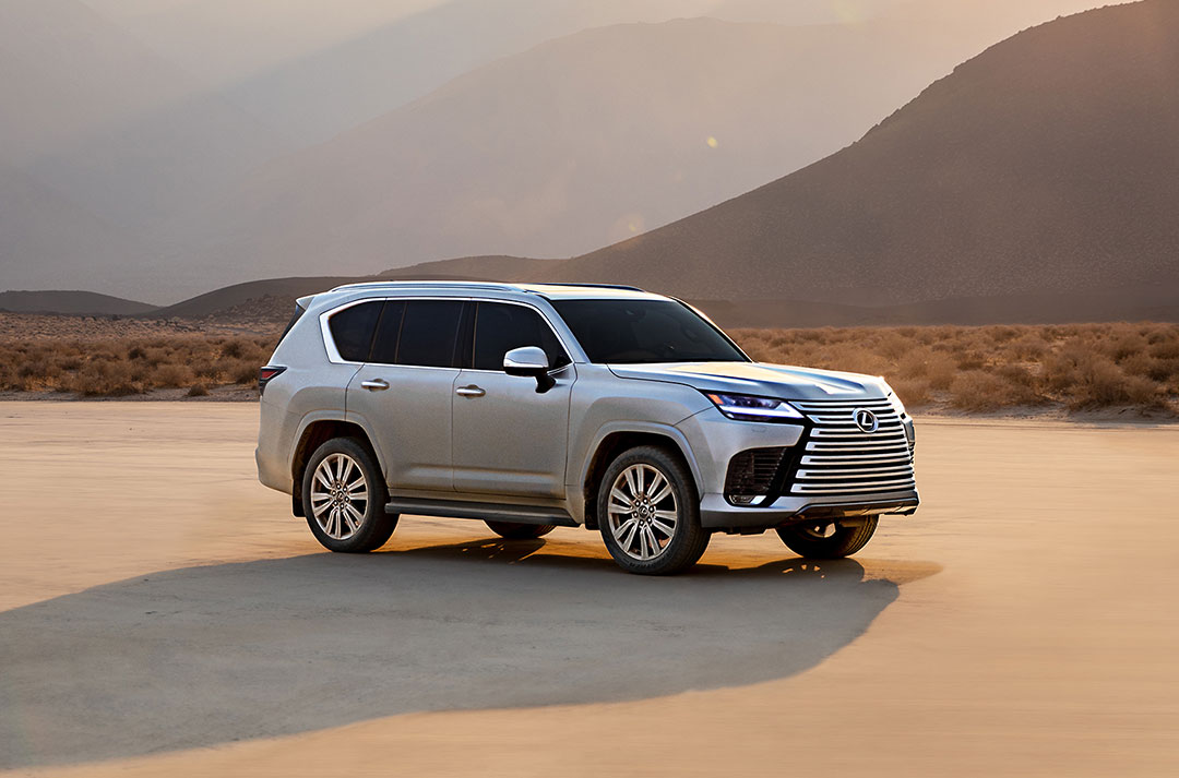 A silver Lexus LX vehicle parked on sandy ground with a mountain in the background