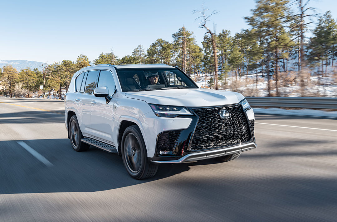 A white Lexus LX SUV drives on a road with trees and mountains in the background
