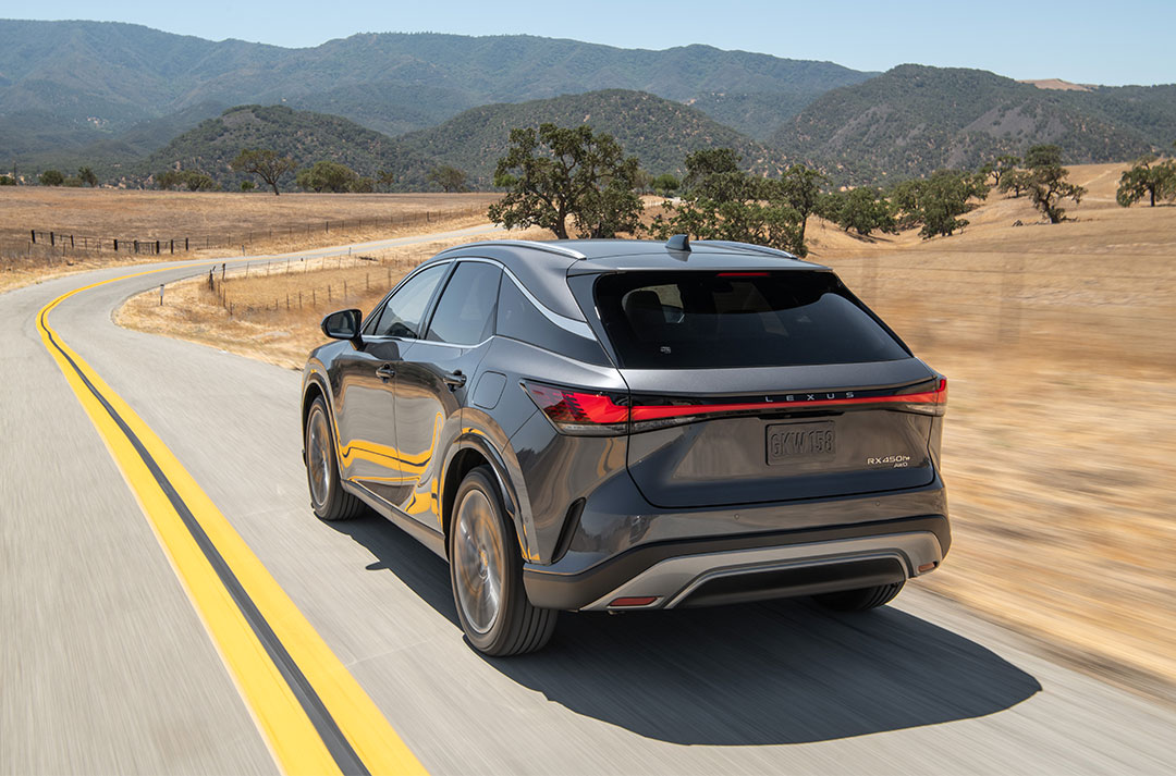A black Lexus RX 450h+ plug-in hybrid vehicle driving along a road with hills in the background
