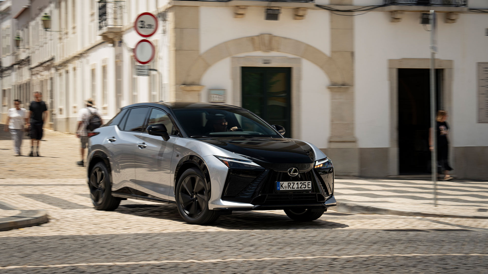 Silver and black Lexus RZ electric SUV driving through a city street with pedestrians and historic buildings.