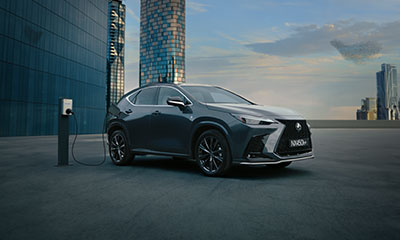 Dark grey Lexus NX 450h+ plug-in hybrid SUV is charging at a public station in a city setting.